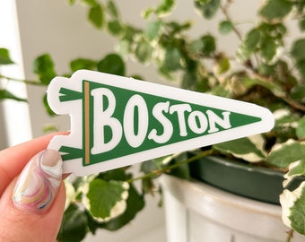May include: A green and white pennant sticker with the word "BOSTON" in white lettering. The sticker is being held by a hand with a colorful nail polish.