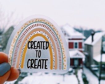 May include: A clear sticker featuring a rainbow design with the words "CREATED TO CREATE" in black. The rainbow has layers of yellow, brown, and pink, with dotted and striped details. The sticker is held up against a snowy outdoor backdrop.