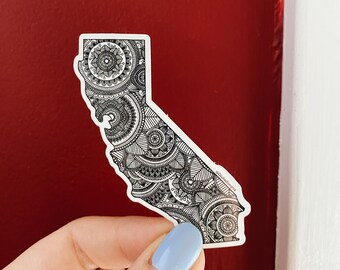 May include: A white sticker in the shape of California, filled with intricate black and white mandala designs. The sticker is held against a red background, showcasing the detailed patterns and the state's outline.