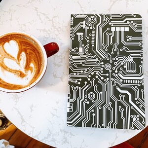 May include: A white ceramic coffee cup with a red handle, filled with coffee and a latte art design. The cup is sitting on a white marble table next to a black and white notebook with a circuit board pattern.