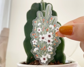 May include: A clear sticker with a white floral bouquet design. The flowers are white with brown centers and are surrounded by green stems and orange berries.