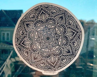 May include: A circular sticker featuring a detailed black and white mandala design. The intricate pattern includes various geometric shapes and floral elements. The sticker is affixed to a window, with a blue sky visible in the background. The artist's name is visible at the bottom.