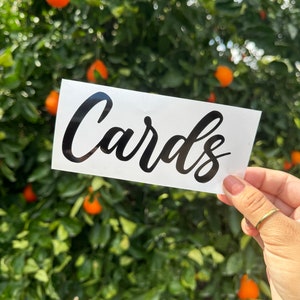 May include: White card with the word "Cards" written in black cursive lettering. The card is held in front of a blurred background of a green tree with orange fruit.