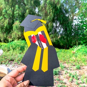 May include: A black graduation cap and gown with a yellow accent and a Sephora gift card inside. The gift card has a red lip design and the text "SEPHORA" on it.