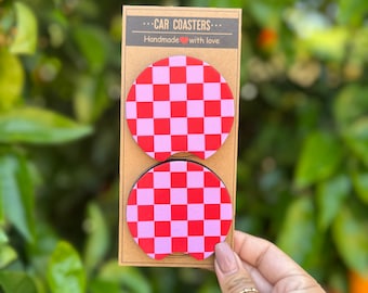 May include: Two car coasters with a red and light pink checkerboard pattern. The coasters are round and are displayed on a brown cardboard backing. The text on the backing reads "CAR COASTERS" and "Handmade with love."