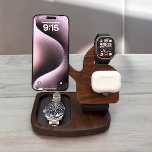 May include: A wooden charging station with a phone stand, watch stand, and a space for wireless earbuds. The phone stand has a black phone with a screen displaying the time "9:15". The watch stand has a black smartwatch with a white face displaying the time "9:15:25". The station is made of dark wood and has a black leather strap.