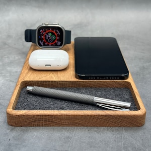 Könnte beinhalten: Eine Holz-Ladestation mit grauer Filz-Auskleidung. Die Station hat drei Steckplätze zum Aufladen eines Smartphones, einer Smartwatch und kabelloser Ohrhörer. Ein Stift ist ebenfalls in der Station enthalten.