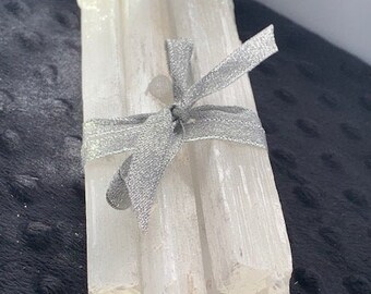 May include: A bundle of four long, translucent white selenite crystal wands with a fibrous, columnar texture. The crystals are held together by a shimmering silver ribbon tied in a bow around the center of the vertical stack of mineral rods.