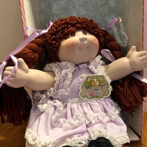 May include: A Cabbage Patch Kids doll with brown yarn hair styled in two braids tied with light purple ribbons. The doll has a soft fabric face, wears a light purple dress with white lace trim, and has black shoes. A branded tag is attached.