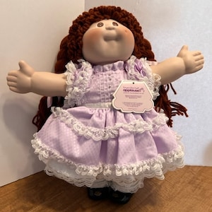 May include: A Cabbage Patch Kids doll with auburn yarn hair, a round face, and rosy cheeks. The doll wears a light purple dress with a white lace-trimmed collar, sleeves, and hem. A white tag labeled Applause is attached to the front of the dress.