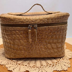 May include: A beige vanity case with a textured crocodile skin pattern. The rectangular bag features a top handle, a zippered closure with two metal pulls, and a structured shape designed for holding personal items or cosmetics securely.