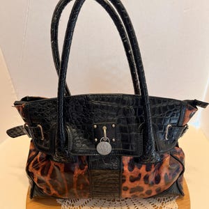 May include: A black handbag with a glossy crocodile-embossed texture on the top panel and handles, featuring a leopard print pattern on the lower section. A silver-toned circular charm with the text NY & C is attached to the center front buckle.
