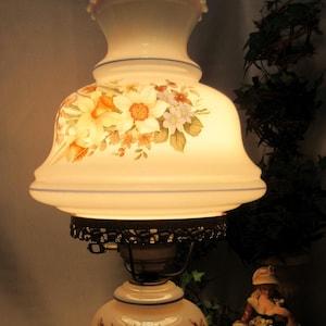 Puede incluir: Una lámpara de vidrio blanco con un diseño floral y una base de metal. La lámpara está encendida y proyecta un brillo cálido. La lámpara está sentada sobre un mantel de encaje blanco.