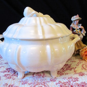 以下が含まれることがあります： 蓋、取っ手、3つの小さな足が付いた白いセラミック製のスープ皿。 スープ皿はリブのデザインで、蓋には装飾的なハンドルが付いています。 背景には置物とバラがあります。