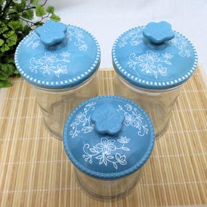 May include: Set of three glass canisters with blue and white floral patterned lids. The lids have a raised flower knob.
