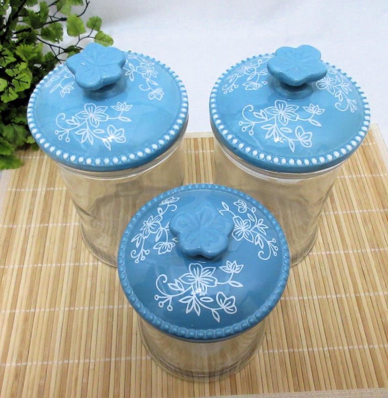 May include: Set of three glass canisters with blue and white floral patterned lids. The lids have a raised flower knob.