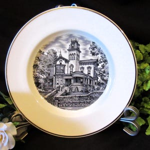Puede incluir: Un plato de cerámica blanco con un borde dorado presenta una ilustración en blanco y negro de una casa de estilo victoriano. La casa tiene una torre alta y un porche con columnas. El plato es una pieza decorativa y sería una gran adición a cualquier hogar.