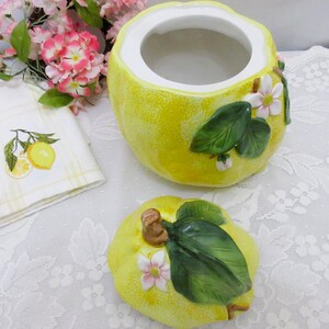 May include: A yellow ceramic jar shaped like a lemon with a matching lid. The jar and lid feature raised, three-dimensional green leaves and small white and pink flowers. The interior of the jar is white, and the exterior has a textured surface.
