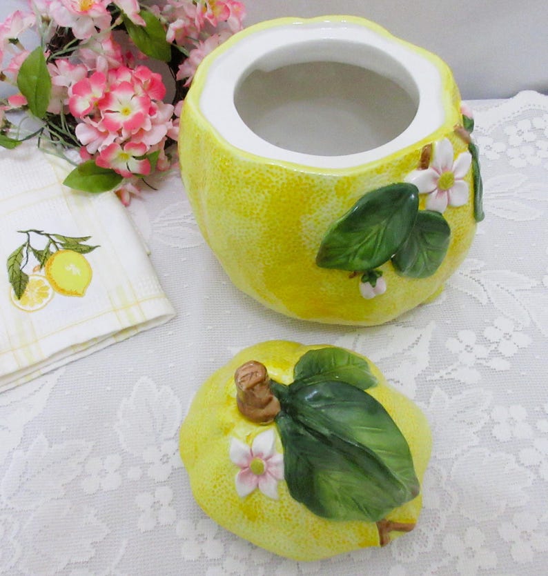 May include: A yellow ceramic jar shaped like a lemon with a matching lid. The jar and lid feature raised, three-dimensional green leaves and small white and pink flowers. The interior of the jar is white, and the exterior has a textured surface.