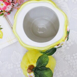 May include: A ceramic lemon-shaped jar with a removable lid. The jar features a textured yellow exterior, a smooth white interior, and a lid adorned with sculpted green leaves, a brown stem, and small white and pink blossoms on the top surface.