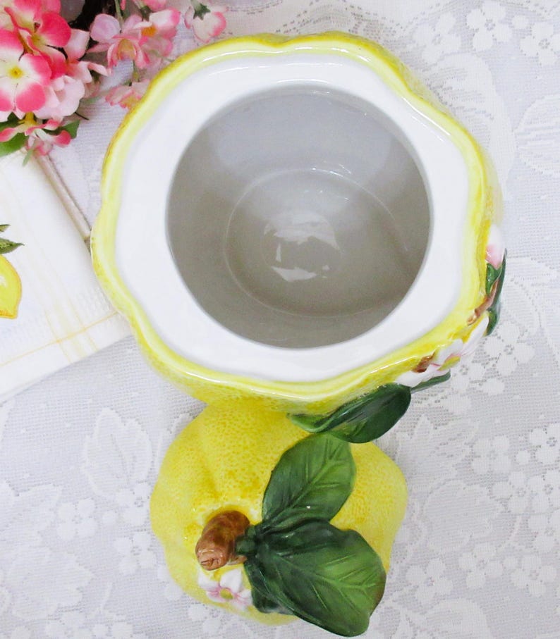 May include: A ceramic lemon-shaped jar with a removable lid. The jar features a textured yellow exterior, a smooth white interior, and a lid adorned with sculpted green leaves, a brown stem, and small white and pink blossoms on the top surface.