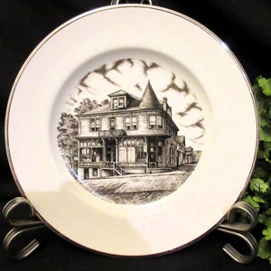 Può includere: Un piatto in ceramica bianca con un'illustrazione in bianco e nero di un edificio con un tetto a punta. Il piatto ha un bordo argentato.