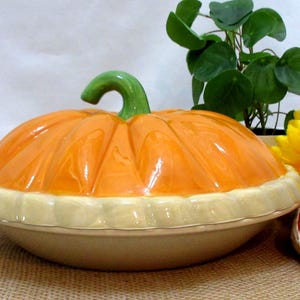 以下が含まれることがあります： 緑色の茎とクリーム色のベースが付いた、カボチャ型のセラミック製サービングディッシュ。オレンジ色の蓋には溝があり、皿はジュートの表面に置かれています。黄色のヒマワリと小さな植物が背景にあります。