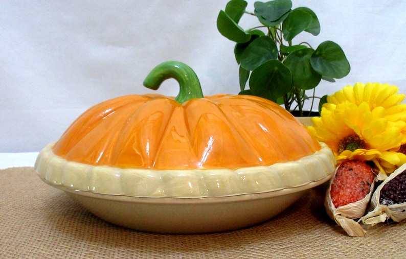 以下が含まれることがあります： 緑色の茎とクリーム色のベースが付いた、カボチャ型のセラミック製サービングディッシュ。オレンジ色の蓋には溝があり、皿はジュートの表面に置かれています。黄色のヒマワリと小さな植物が背景にあります。