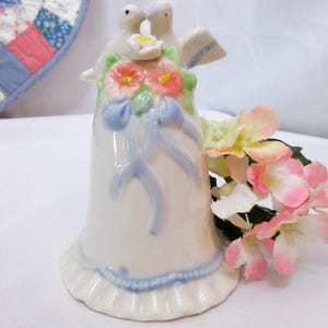 Op de afbeelding: Een witte keramische bruidsbel met twee kussende duiven aan de bovenkant. De bel is versierd met een blauw lint en bloemmotieven. Een boeket roze en gele bloemen ligt ernaast.