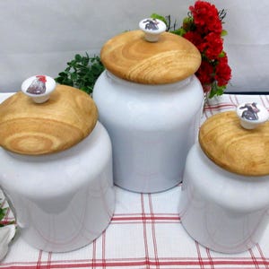 Könnte beinhalten: Drei weiße Keramikbehälter mit Holzdeckeln. Jeder Deckel hat einen dekorativen Keramikknopf mit einem Hahn, einer Kuh oder einem Schwein. Die Behälter stehen auf einer rot-weiß karierten Tischdecke.