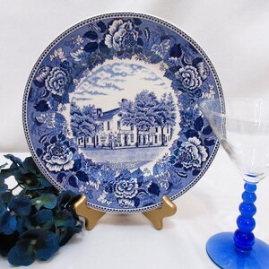 Peut inclure: Une assiette décorative bleue et blanche avec une bordure florale et une scène centrale d'un bâtiment. L'assiette est présentée sur un support doré, avec un verre à martini à tige bleue à droite et des fleurs bleues à gauche.