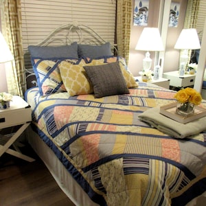 Pode incluir: Uma cama com cabeceira de metal branco é coberta com um edredom de patchwork em tons de azul, amarelo e pêssego. Almofadas decorativas em azul, amarelo e cinza são dispostas na cama. Uma pequena bandeja com rosas amarelas está em cima de um cobertor dobrado.