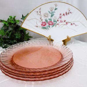 May include: A stack of four pink glass plates with a textured, wavy rim. A decorative white fan with a floral design and gold trim is in the background. The plates are approximately 8 inches in diameter.