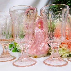 Könnte beinhalten: Sechs rosafarbene Weingläser mit geätzten floralen Mustern. Die Gläser mit abgerundeten Böden sind in einer Reihe angeordnet. Eine dekorative Figur und Blumen sind im Hintergrund zu sehen.
