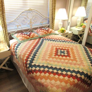 Peut inclure: Un couvre-lit avec un motif en patchwork en forme de losange dans des tons orange, rouge, bleu et crème. Le couvre-lit est composé de petits morceaux de tissu carrés disposés en dégradé, recouvrant un lit avec une tête de lit en métal blanc.