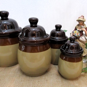 Pode incluir: Conjunto de quatro potes de cerâmica marrom e bege com tampas. Os potes são de tamanhos diferentes e têm um acabamento esmaltado.