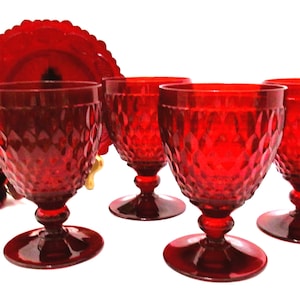 May include: A set of four ruby red glass goblets featuring a diamond-patterned texture on the bowls and a circular base. Each goblet has a short, thick stem. A red glass plate with a scalloped edge and two red fabric roses are visible behind.