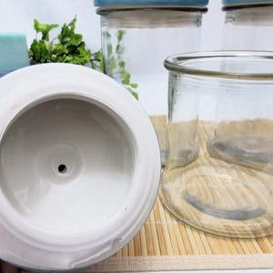 May include: A white ceramic lid with a small hole in the centre. The lid is sitting on top of a wooden surface. Three clear glass jars with blue lids are visible in the background.