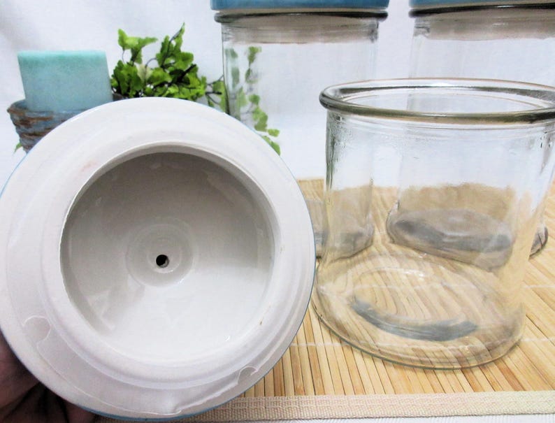 May include: A white ceramic lid with a small hole in the centre. The lid is sitting on top of a wooden surface. Three clear glass jars with blue lids are visible in the background.