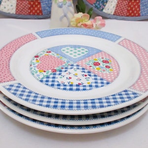 以下が含まれることがあります： パッチワークデザインの白いセラミックプレート3枚のスタック。プレートにはピンクのドット、青のギンガムチェック、花柄が施されています。プレートは積み重ねられています。
