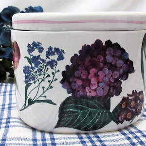 Op de afbeelding: Witte keramische pot met een bloemenpatroon. De pot heeft een roze deksel en een bloemenpatroon met paarse, blauwe en groene bloemen.