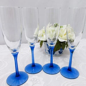 May include: Four clear champagne flutes with bright blue stems and bases. The glasses are arranged in a row, highlighting their elegant design. The blue colour provides a striking contrast to the clear glass, perfect for celebrations.