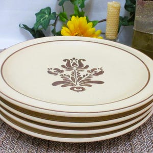 May include: A stack of four beige ceramic plates with a brown floral design in the center. Each plate has a brown rim and a brown border. The plates are stacked on top of each other.