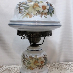 Peut inclure: Une lampe en porcelaine blanche avec un motif floral. La lampe a un abat-jour blanc avec une bordure bleue et une base dorée.