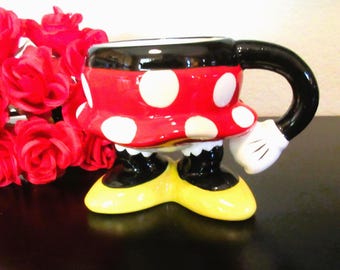 May include: A ceramic mug shaped like a cartoon character, featuring a red body with white polka dots, a black rim, and a black handle ending in a white gloved hand. The base consists of two black legs wearing yellow shoes, providing a stable stand.