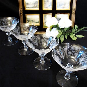 Könnte beinhalten: Vier klare Champagner-Coupe-Gläser mit geätzten floralen Designs. Die Gläser haben eine breite, flache Schale und einen langen Stiel. Die Gläser sind auf einer schwarzen Oberfläche angeordnet, mit einem gerahmten Kunstwerk und weißen Blumen im Hintergrund.