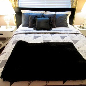 May include: A bed with a black headboard, white bedside tables, and a patterned quilt. The bed is made with dark blue pillows and a black fluffy throw blanket. Two lamps with white shades sit on the bedside tables.