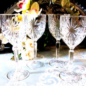 May include: A set of four clear glass wine goblets featuring a prominent starburst or sunburst cut pattern on the bowl. Each glass has a faceted, multi-sided stem and a circular base, reflecting light through the intricate, etched crystal design.