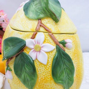 May include: A ceramic lemon-shaped jar with a removable lid. The yellow textured surface features raised, hand-painted details including green leaves, brown branches, and white blossoms with yellow centers. The lid is topped with a green leaf.