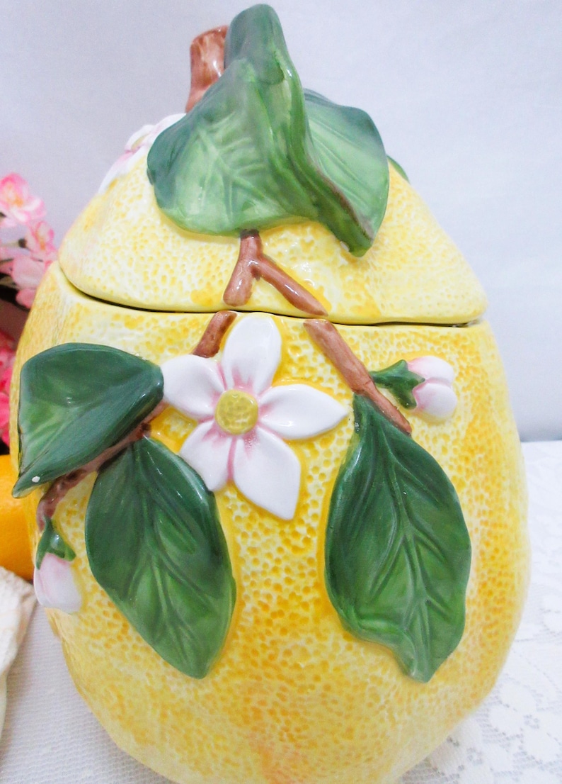 May include: A ceramic lemon-shaped jar with a removable lid. The yellow textured surface features raised, hand-painted details including green leaves, brown branches, and white blossoms with yellow centers. The lid is topped with a green leaf.